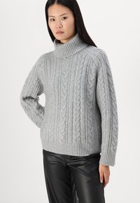 Pull en maille grise avec un col roulé haut, un design en tricot à torsades, des poignets et un ourlet côtelés ; porté avec un pantalon en cuir noir.