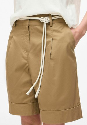 Vyras dėvi smėlio spalvos šortus iki kelių su klostėmis ir baltą virvinę diržą, surištą mazgu, ranka kišenėje, iš dalies matoma balta marškinėliai.