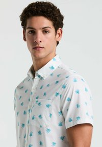Korte mouwen witte shirt met turkooise bladerenpatroon, button-down kraag en linkerkant borstzak. Gemaakt van lichtgewicht stof.