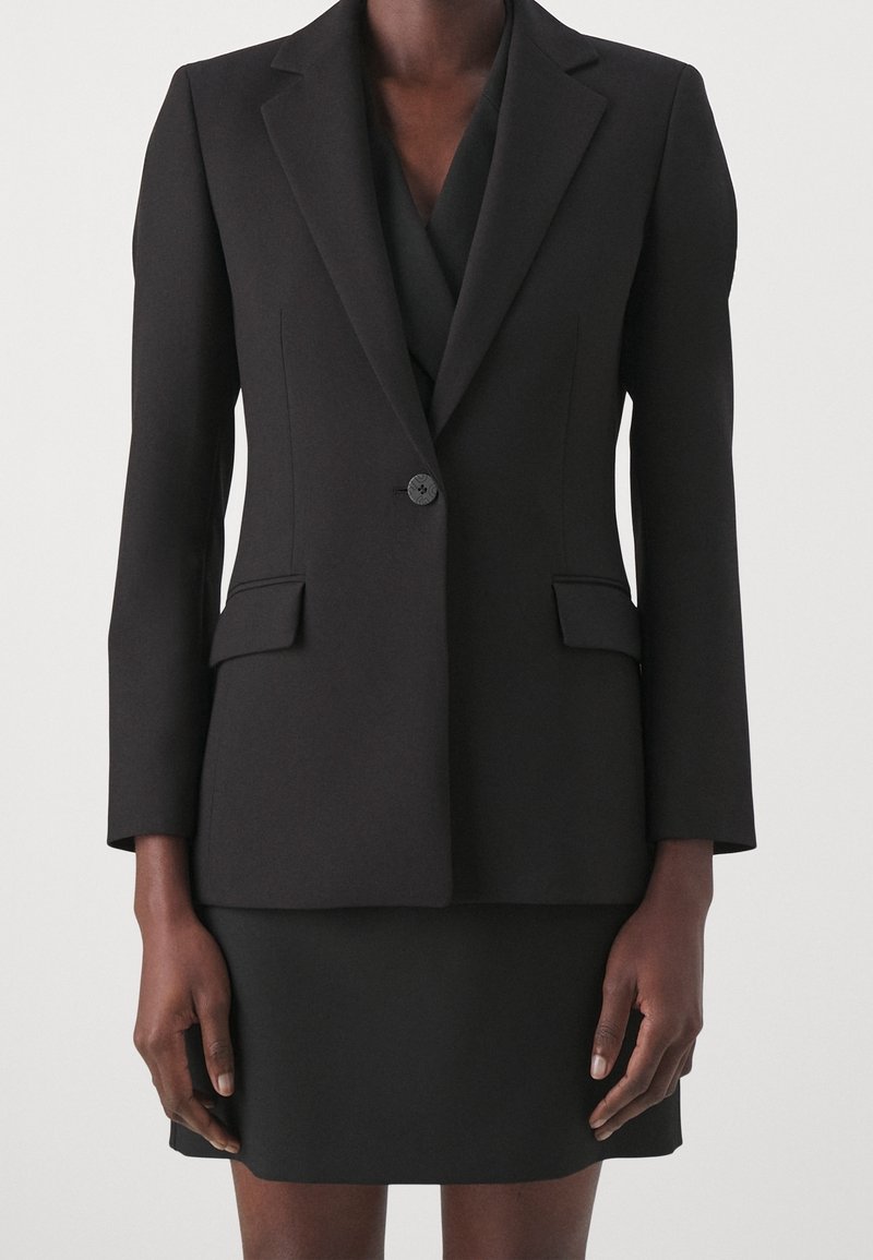 Veste noire cintrée avec une fermeture à un bouton, revers crantés, poches à rabat, portée avec une jupe noire assortie.