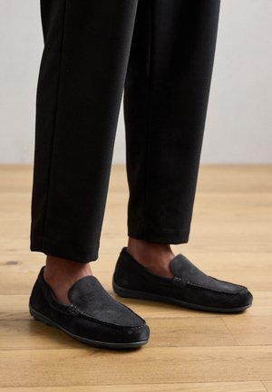 Mustad nubukist slip-on kingad, millel on ümar nina ja õmblusdetailid. Ühendatud mustade pükstega, mille taustal on puidust põrand.