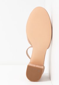Sandale à talon en daim nude avec une semelle lisse en caoutchouc beige. Elle possède une fine bride à la cheville et un talon carré pour le maintien. Surface inférieure plate.