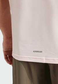 T-shirt texturé rose clair avec un col rond, arborant un logo noir "AEROREADY" à l'ourlet. Couture subtile sur le bord inférieur.