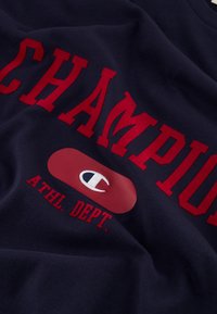 Marineblauwe katoenen sweatshirt met in rode letters "CHAMPION" en een rode ovaal met een wit logo, met gelaagde, verheven letters voor textuur.