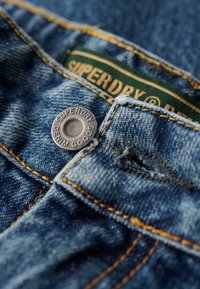 Jeans dżinsowe w ciemnoniebieskim kolorze. Posiadają srebrny metalowy guzik z wygrawerowanym napisem "SUPERDRY DENIM GOODS" oraz kontrastowe żółte szwy.