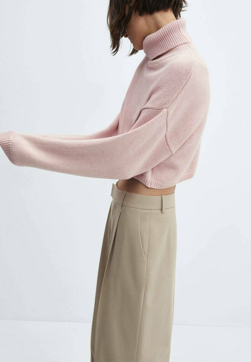 Helles rosa, kurz geschnittenes Sweatshirt mit hohem Rippenkragen, das strukturierter Strickstoff und lockere Ärmel zeigt, kombiniert mit beigefarbenen, hochgeschnittenen Hosen.