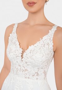 Abito bianco con un corpetto in pizzo floreale, scollatura a V e spalline larghe, che si apre in una gonna fluida. Il pizzo presenta dettagli intricati.