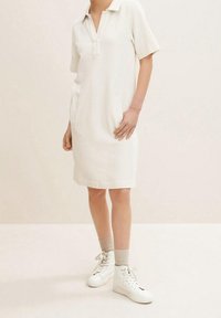 Kurzarm-Wickelkleid aus hellem Beigefarbigen Stoff, mit zwei seitlichen Taschen, gerade geschnitten und kombiniert mit weißen High-Top-Sneakern und beigen Socken.