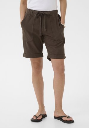 Donna che indossa pantaloncini con coulisse di colore marrone scuro con orli arrotolati e infradito neri, in piedi contro uno sfondo bianco.