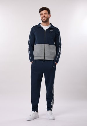 Man in navy and grey tracksuit with white sneakers, standing with hands in pockets, smiling and looking slightly to the side.