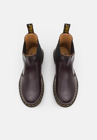 Par de botas Chelsea de cuero marrón oscuro con costuras amarillas y paneles elásticos negros, vistas desde arriba sobre un fondo blanco.