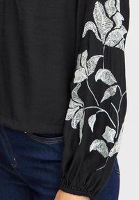 Blouse noire à manches longues ornée de broderies en forme de feuilles argentées. Le tissu a une texture douce, et les manches sont froncées au niveau des poignets.