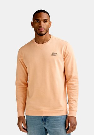 Mand iført ferskenfarvet langærmet sweatshirt med lille sort logo på brystet og blå jeans, stående mod en ensfarvet lys baggrund.