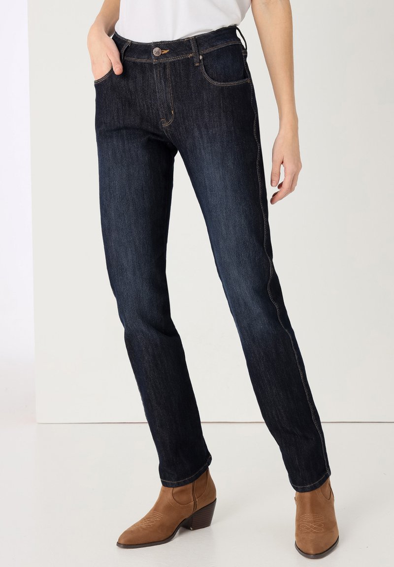 Jeans de mezclilla azul oscuro con diseño de pierna recta, cinco bolsillos y cierre de botón. Llevados con botas de tobillo de tacón marrones.