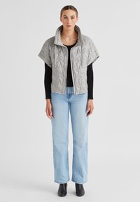 Grau gesteppte Jacke mit hohem Kragen und kurzen Ärmeln, mit Reißverschluss vorne, kombiniert mit schwarzem Langarmoberteil und hellblauen Jeans.
