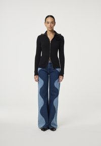 Zwarte ribgebreide ritstrui met een kraag en bloemaccent, gecombineerd met licht en donkerblauwe flared jeans met golvende naden.