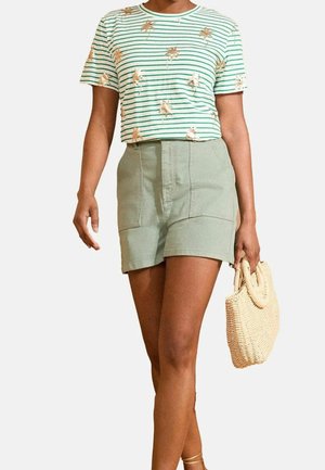 Femme portant un t-shirt rayé vert avec un motif de palmier, un short taille haute vert clair, et tenant un sac en paille tressée.