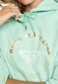 Hoodie v mátově zelené barvě s provázkem na stažení a zlatým potiskem s nápisem "ROXY SINCE 1990" a logem srdce. Měkká textilie.
