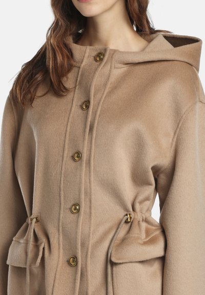 Palton bej cu glugă, confecționat dintr-un material moale, prevăzut cu nasturi aurii, talie cu sfoară și buzunare frontale cu clapetă. Textură netedă și design casual.