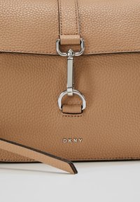 Bolso de piel marrón con textura de guijarro, tapa plana, cierre de hardware plateado y logo "DKNY" embossado en la parte frontal.