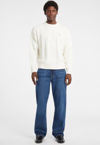Maglione bianco a treccia con collo rotondo e orlo a costine, abbinato a jeans blu scuro a vestibilità ampia e scarpe nere, in piedi davanti a uno sfondo neutro.