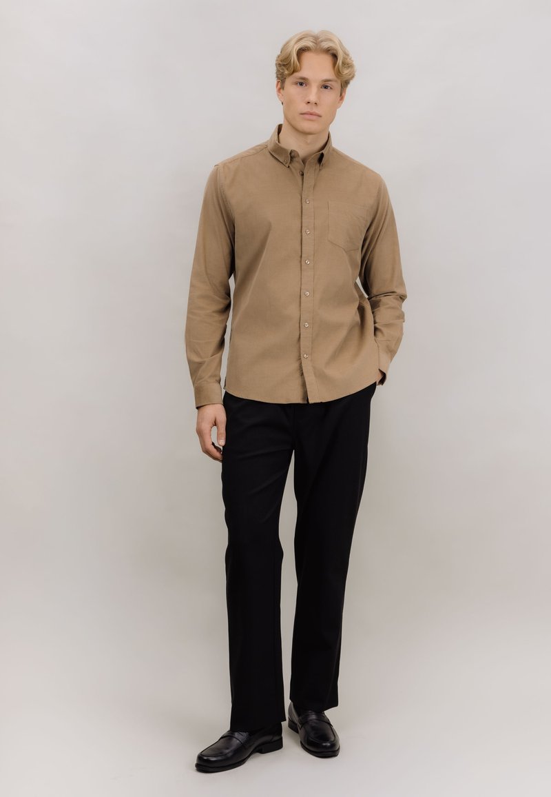 Langermet skjorte med knapper i lyst brunt stoff med brystlomme, kombinert med svarte bukser og svarte sko.