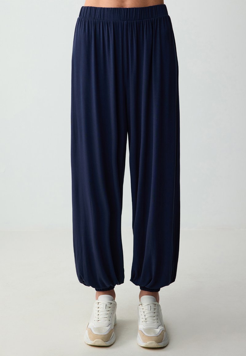 Pantaloni navy blue con vita elastica, dalla vestibilità ampia e tapered e caviglie arricciate, abbinati a scarpe sportive bianche. Tessuto dalla struttura liscia.