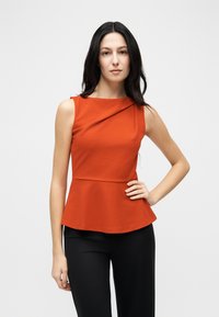 Γυναίκα με μακριά σκούρα μαλλιά, φορώντας αμάνικο πορτοκαλί peplum τοπ και μαύρο παντελόνι, στάση με το ένα χέρι στη μέση σε απλό φόντο.