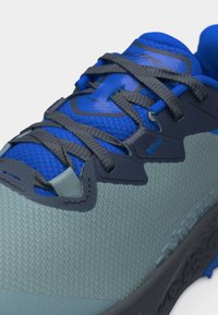 Κοντινή λήψη αθλητικού παπουτσιού σε μπλε και γκρι χρώμα που δείχνει υφασμάτινη υφή, γκρι κορδόνια και ετικέτα "TIMP 6" στη γλώσσα.