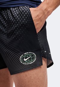 Gros plan d'une personne portant un short de course Nike noir avec un motif géométrique et un logo Nike près de l'ourlet, main dans la poche.