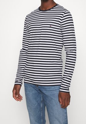 Homme portant une chemise rayée noire et blanche à manches longues, associée à un jean en denim bleu, se tenant devant un fond uni.