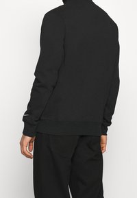 Black sweatshirt with a high collar, ribbed cuffs, and waistband. Features a subtle logo accent on the sleeve. Soft, jersey fabric.