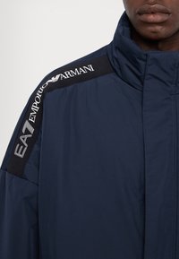 Tengerészkék, párnázott dzseki magas gallérral. Jellemzője a vállon található fekete szövetpanel fehér "EA7 Emporio Armani" logóval. Simaság jellemzi.