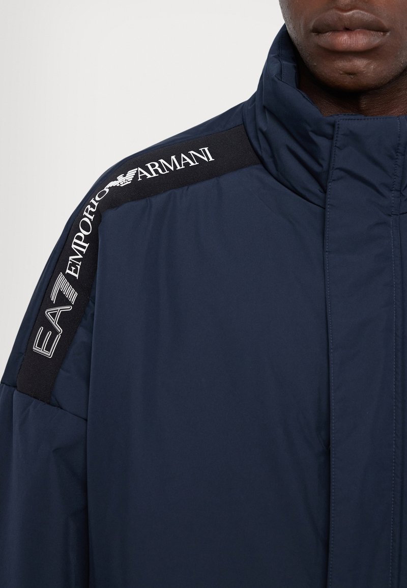 Chaqueta acolchada de color azul marino con cuello alto. Presenta un panel de tela negra en el hombro con el logo blanco "EA7 Emporio Armani". Textura suave.