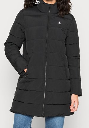 Zwarte doorgestikte lange winterjas met een ritssluiting aan de voorkant, twee ritssluitzakken en een wit "cK"-logo op de borst, gedragen over een blauwe spijkerbroek.