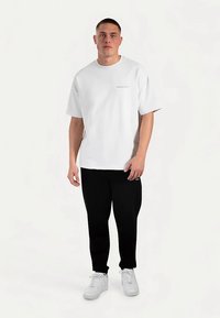 T-shirt en coton blanc à manches courtes, avec un petit logo beige sur la poitrine. Associé à un pantalon noir et des baskets blanches.