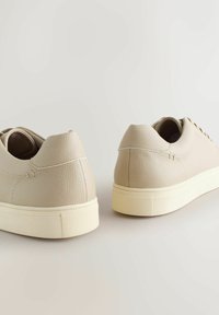 Beige leather sneakers with a textured surface, rounded heel, and contrasting light rubber sole. Shoelace holes and minimal stitching detail.