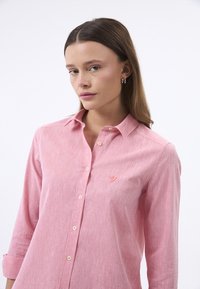 Lys pink skjorte med krave, der har et struktureret stof og en diskret broderet logo på venstre bryst. Klassisk pasform med lange ærmer.
