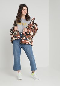 Kamikazi pūkų striukė žemės tonų, derinama su pilku marškinėliais ir mėlynomis trumpomis džinsomis. Baltos sportinės avalynės su neoninio geltono akcentais derinys užbaigia aprangą.