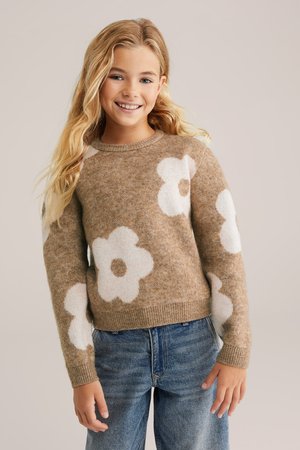 Helle braune Strickpullover mit großen weißen Blumenmustern, runder Halsauschnitt und gerippten Bündchen, kombiniert mit blauen Jeans.