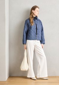 Quilted blue jacket with a buttoned front, worn over a dark top, paired with white wide-leg pants and a beige bag. Flat beige shoes complete the look.