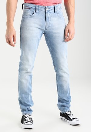 Hombre con vaqueros ajustados azul claro, zapatillas negras y blancas con cordones, y una camisa roja, de pie frente a un fondo liso.