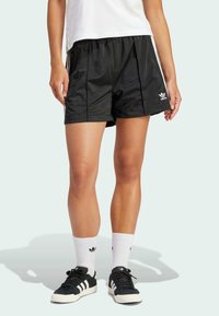 Černé sportovní šortky z hladké látky, s bočním zipem, volným střihem a bílým logem Adidas. Nošeny s černými teniskami a bílými ponožkami.