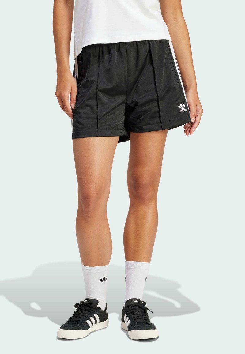 Černé sportovní šortky z hladké látky, s bočním zipem, volným střihem a bílým logem Adidas. Nošeny s černými teniskami a bílými ponožkami.