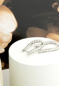 Orecchini a cerchio in argento con un design curvo e una fila di pietre trasparenti sul fronte, esposti su un piedistallo bianco circolare.