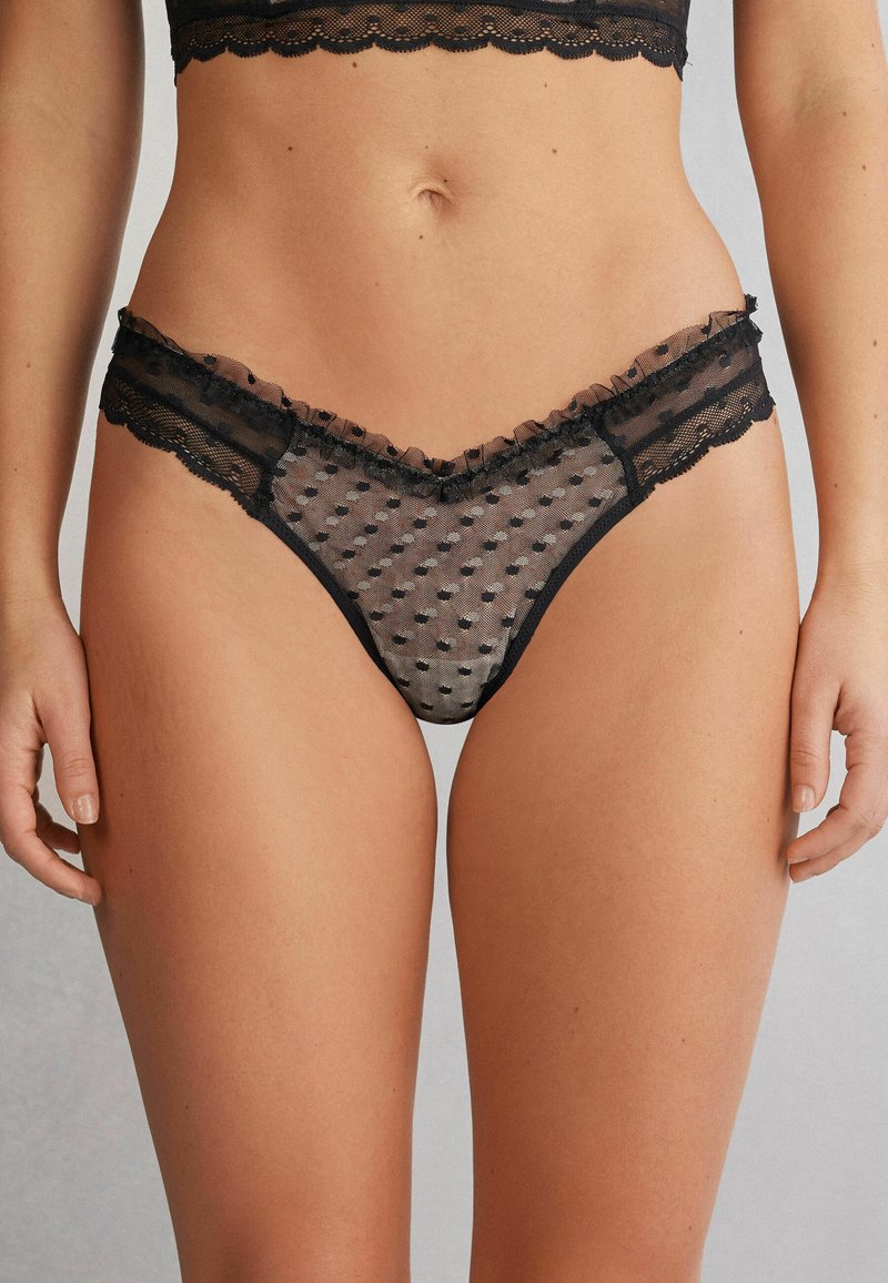 Zwarte kant string met doorschijnend stippenpatroon, gerimpelde rand langs de tailleband en golvende randen. Zachte textuur, lichtgewicht ontwerp.