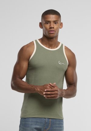 Muž s vypracovanou postavou, oblečený v textúrovanom olivovozelenom tielku bez rukávov s bielym lemom, stojaci so založenými rukami na jednoliatom sivom pozadí.