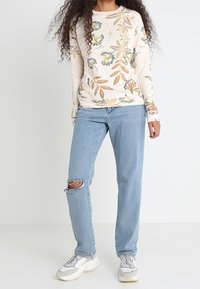 Helle Bluse mit floralem Muster und langen Ärmeln; hellblaue, ausgewaschene Jeans mit einem Riss am Knie; weiße und graue Turnschuhe.