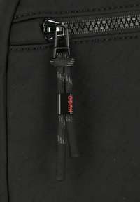 Tira de cremallera negra con un agarre de cordón texturizado, que presenta un logotipo rojo "HUGO" en un acento cilíndrico, adjunto a una superficie de tela negra.