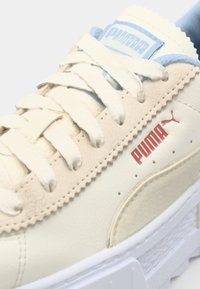 Sneaker mit cremefarbener synthetischer Oberseite, akzentuiert mit hellblauen Elementen, strukturierten Schnürdetails und rotem Puma-Logo an der Seite. Weiße Plateau-Sohle.